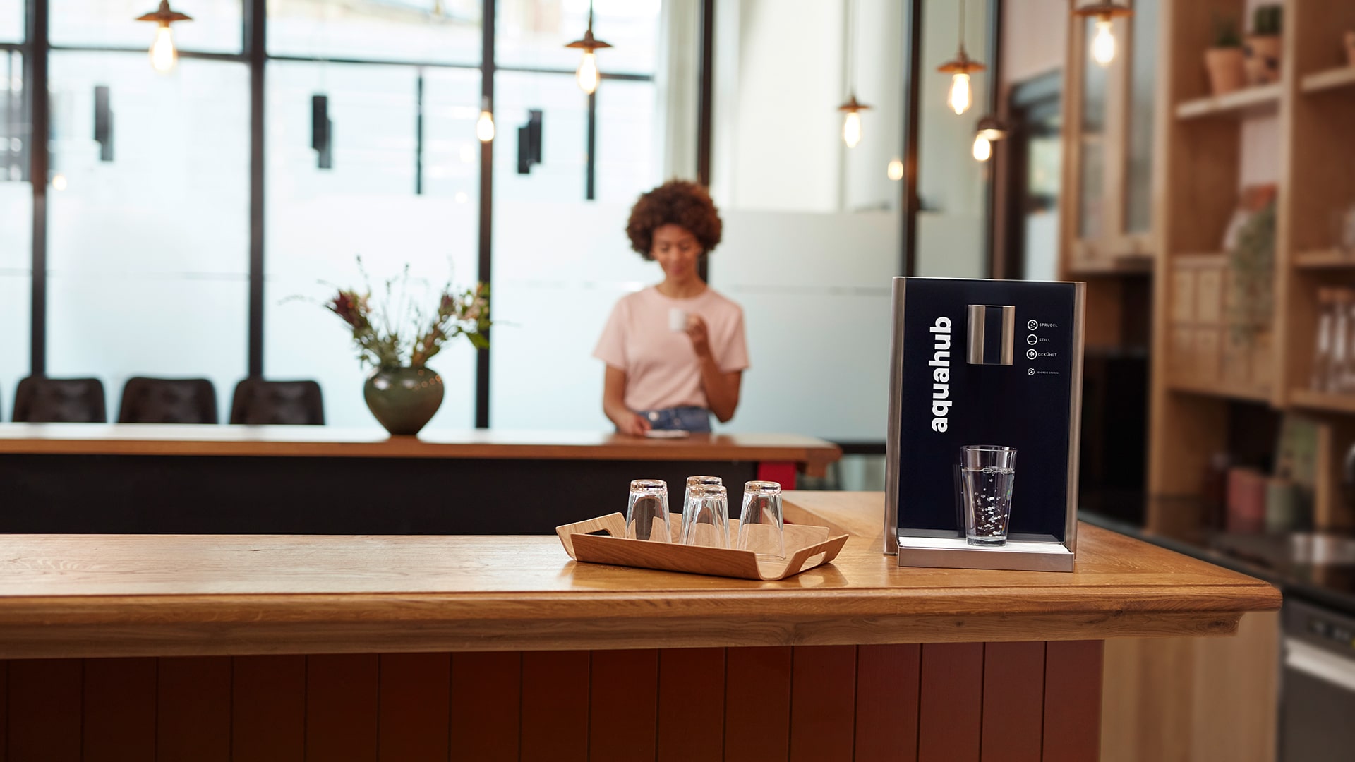 Im Vordergrund steht ein Tablett mit Umgekehrten Gläsern. Rechts daneben steht der Wasserautomat Aquahub. Im Hintergrund steht eine Frau mit einem Kaffeebecher.