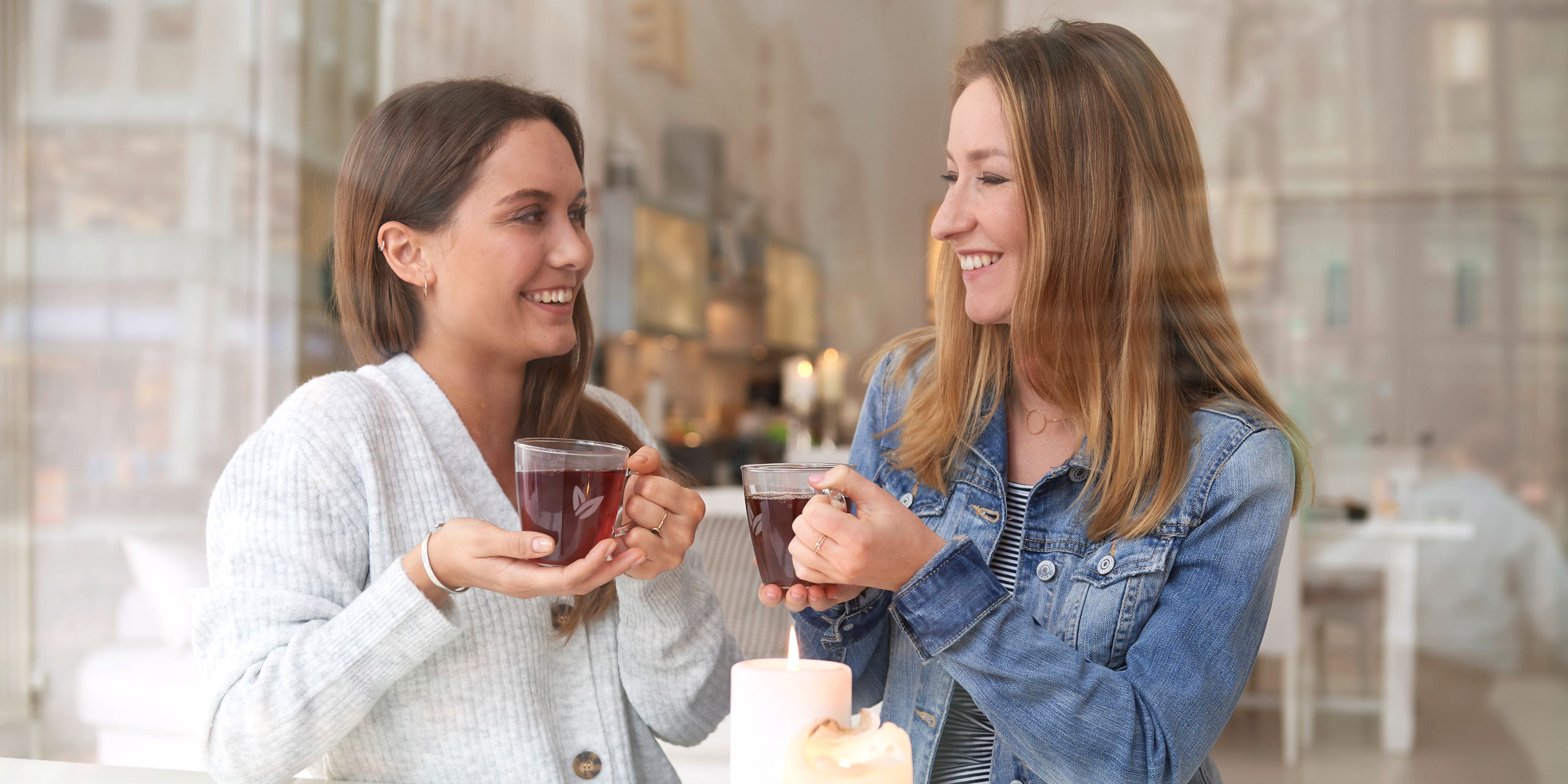 Zwei Frauen hinter einer Glas Wand. Die rechte Frau schaut in die Kamera und die linke Frau schaut weg. Beide trinken Tee. 